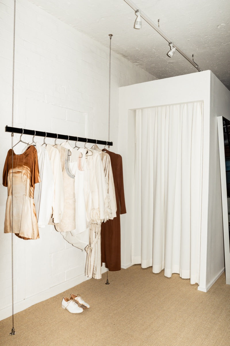 Cream archive garments on a steel rail beside the studio fitting-room curtain.